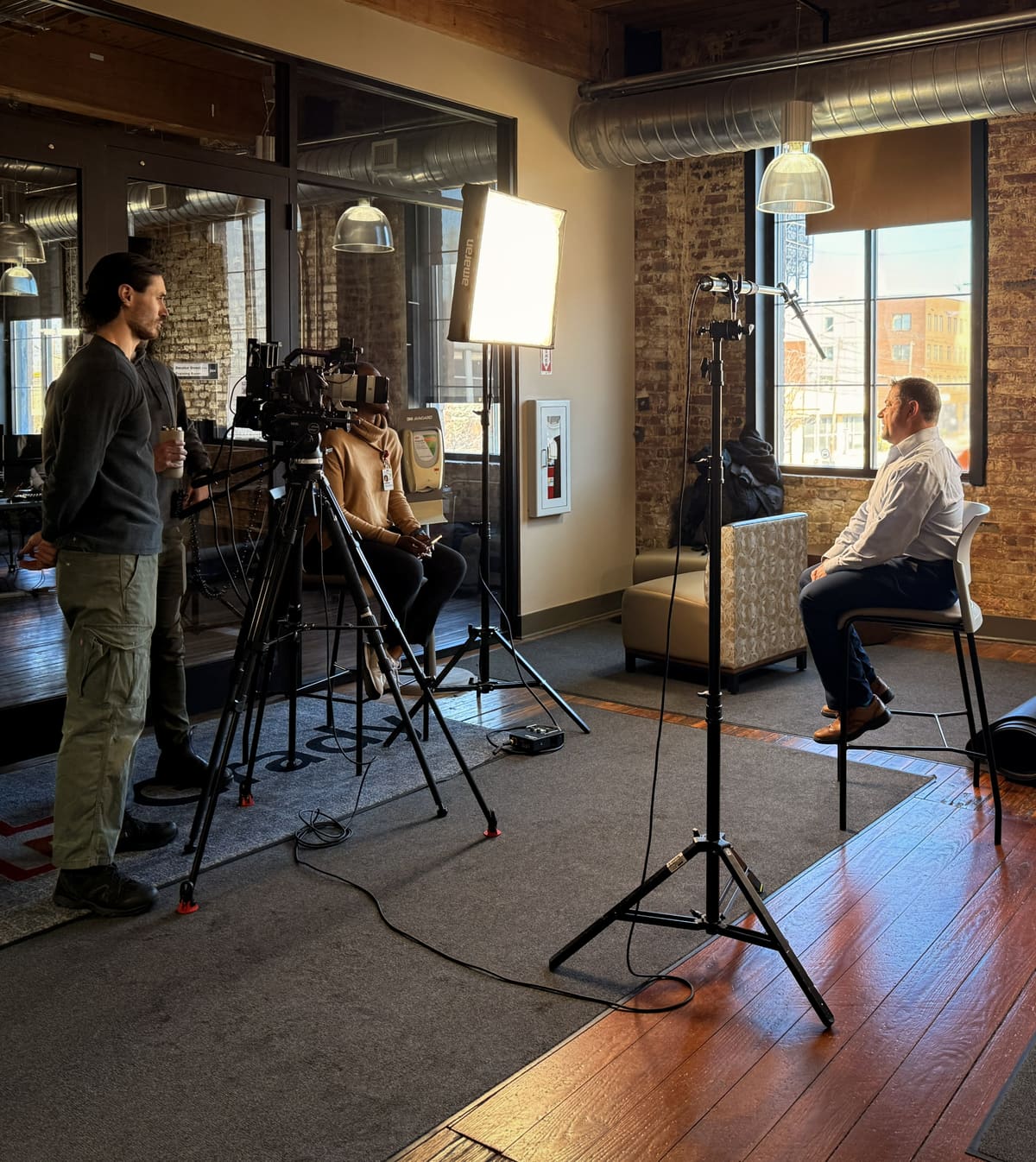 A corporate interview under way on a client's floor: a camera on a tripod, a large soft light on the subject seated by the windows.