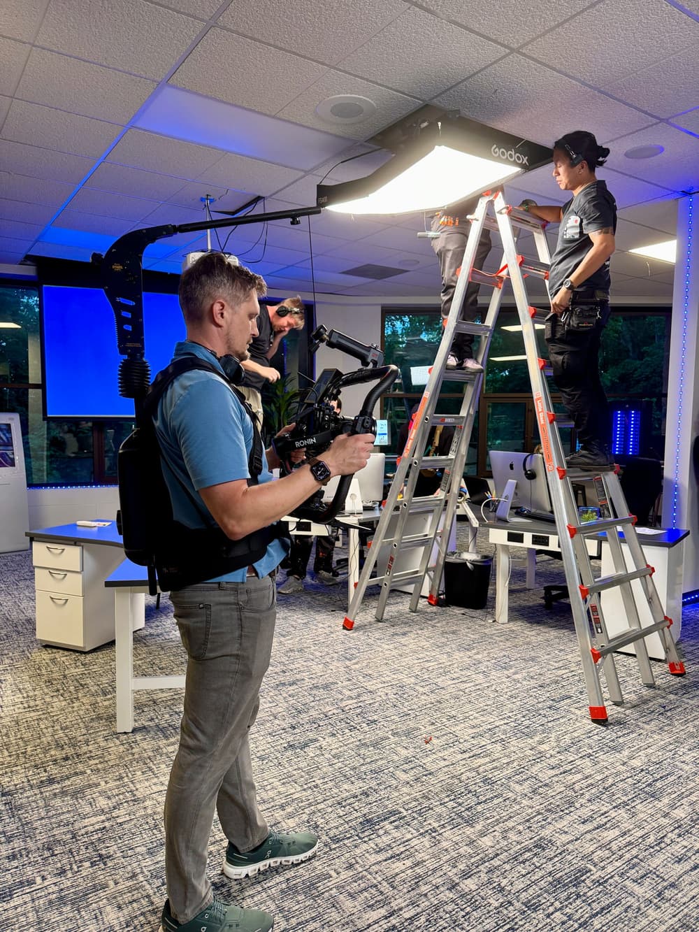 The camera operator on a body-mounted stabilizer while a second crew member on a ladder adjusts a large soft light rigged overhead, in an office lit with blue accent light.