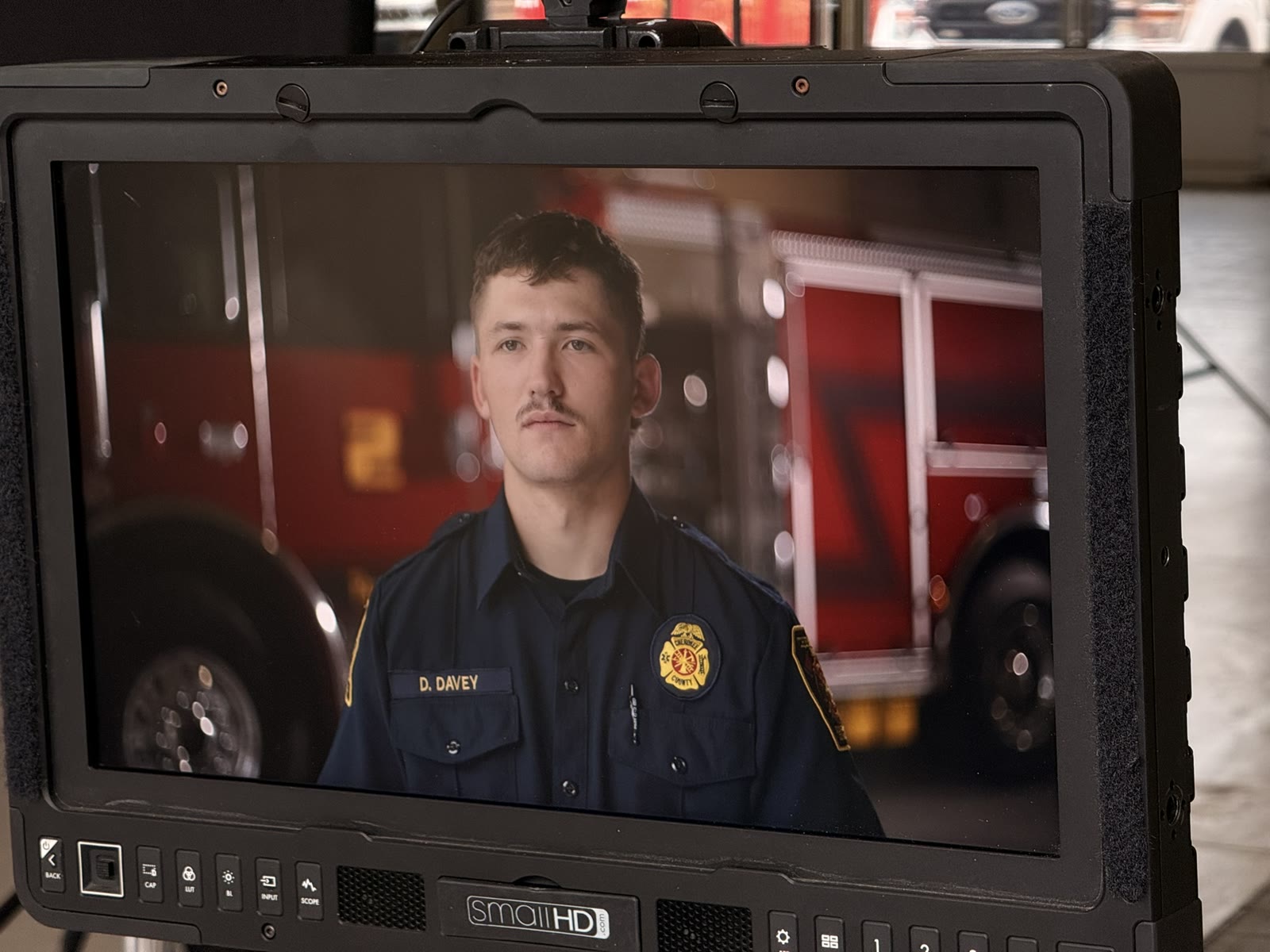 The production monitor showing a framed interview of a firefighter in uniform, a fire engine soft in the background