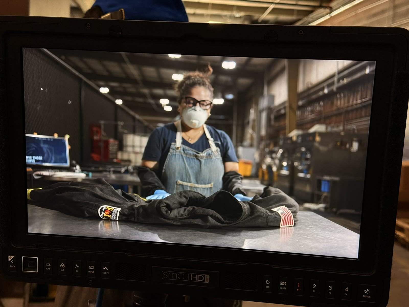 The production monitor showing a masked worker in an apron inspecting protective gear on a steel table in the plant