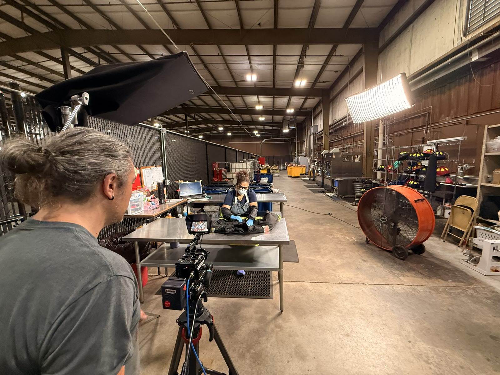 Filming on the plant floor: the camera operator at the tripod, a worker laying out protective gear on a steel table, and a large soft light raised over the scene