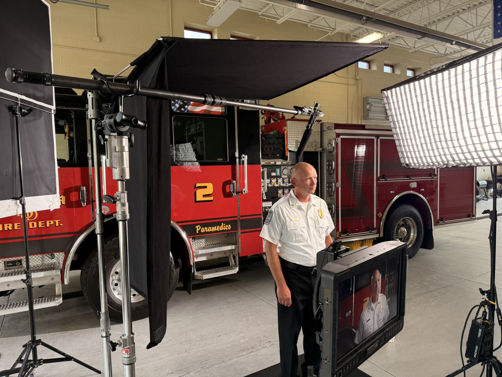 An interview setup inside a fire station: a large soft light and a black overhead shade rigged beside a fire engine, a boom mic overhead, and the fire chief standing by the monitor