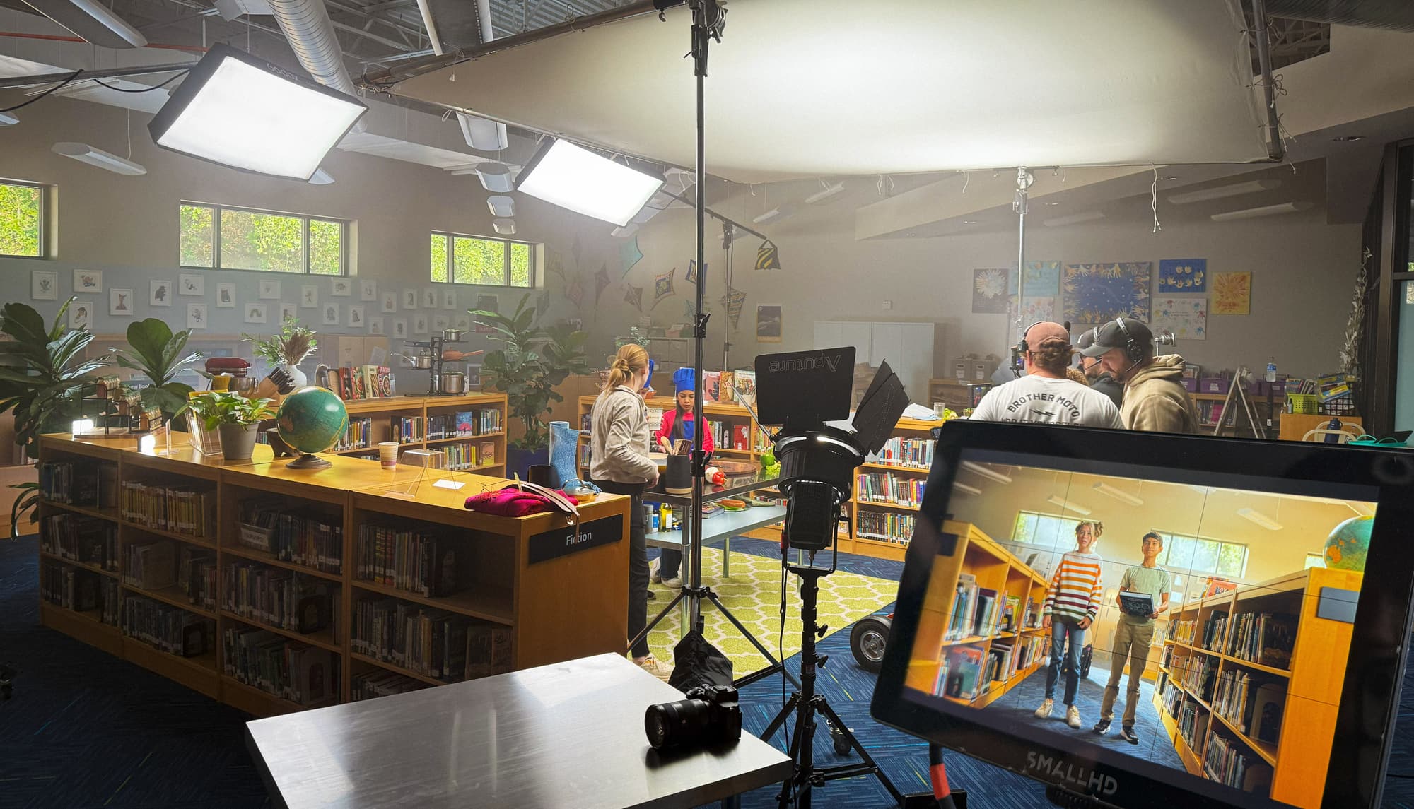 A lighting build on a library location: large LED soft panels rigged overhead, an Aputure fixture with barn doors on a stand, a bounce spanning the ceiling, haze catching the beams, and the framed shot on a monitor where the crew is watching.